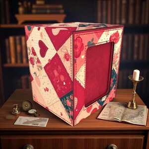 Puede incluir: Una caja decorativa con un diseño de patchwork con corazones y rosas en tonos de rojo, rosa y blanco. La caja tiene una abertura rectangular y está colocada sobre una superficie de madera. Una vela y un mapa también están en la superficie.