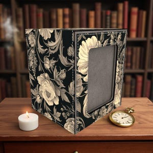 May include: A black and cream floral patterned picture frame with a rectangular aperture. The frame is on a wooden surface with a lit candle and a gold pocket watch. A bookshelf is in the background.