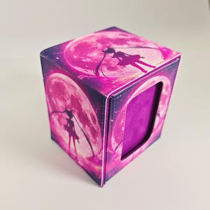 May include: A rectangular deck box featuring a vibrant magenta and purple color scheme with a graphic of a silhouette standing before a large full moon on the top and side panels. The box has a textured exterior and a deep purple interior lining.