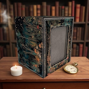 May include: A decorative box with a forest-themed design, featuring a dark teal and black color palette with accents of orange and white. The box has a rectangular frame and a stitched border. A lit candle and a gold pocket watch are on the wooden surface.