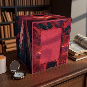 May include: A decorative tissue box with a vibrant red and dark blue abstract design. The box has a rectangular opening and is placed on a wooden surface. A small pocket watch and candle are nearby.