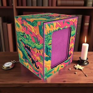 May include: A colorful box with a vibrant design featuring a green alligator, pink, yellow, and orange tropical foliage. The box has a purple interior and a stitched border. A gold pocket watch and a lit candle are nearby.