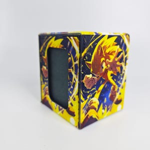 May include: A square-shaped storage box featuring a vibrant graphic of the character Sonic the Hedgehog in a dynamic pose. The design uses bright yellow and blue colors with swirling energy effects. The box has a rectangular cutout on the front side.