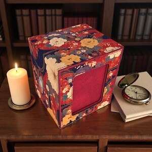 Può includere: Una scatola decorativa con interno rosso e un motivo floreale rosso, giallo e bianco. Una candela bianca accesa e un orologio da tasca su un libro sono anch'essi presenti. La scatola è su una superficie di legno.
