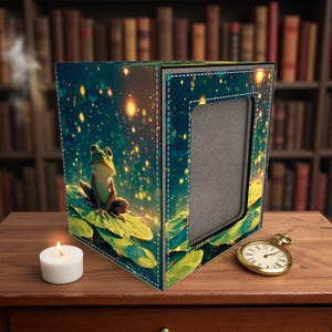May include: A decorative box with a whimsical frog illustration on a lily pad, set against a starry night backdrop. The box has a rectangular opening and is accented with white stitching. A lit candle and a gold pocket watch are on the wooden surface.