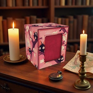 Può includere: Una scatola quadrata rosa con un'apertura incorniciata, decorata con illustrazioni di gatti neri e rami floreali. Due candele accese e una bussola sono su un tavolo di legno, con una mappa e una libreria sullo sfondo.