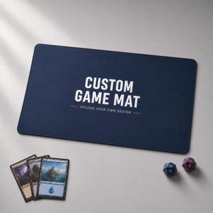 Op de afbeelding: Een rechthoekige donkerblauwe speelmat met de witte tekst CUSTOM GAME MAT en UPLOAD YOUR OWN DESIGN. Drie ruilkaarten zijn linksonder geplaatst en twee veelzijdige dobbelstenen liggen rechtsonder op de mat.