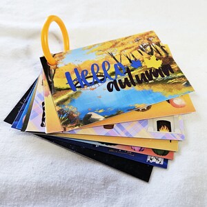 May include: A collection of illustrated cards held together by an orange ring. The top card displays a landscape with a lake and the words "Hello autumn" in blue. Other cards feature autumn-themed illustrations, including a fireplace and pumpkins.