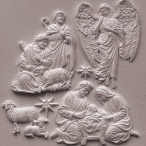 May include: A white relief sculpture featuring a nativity scene with a central manger, figures of a parent and child, a standing figure, a shepherd with a sheep, a winged angel, two additional sheep, and two eight-pointed stars in a matte finish.