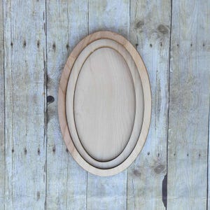 Könnte beinhalten: Ein Satz von drei ovalen Holzrahmen, die ineinander verschachtelt sind. Die Rahmen bestehen aus hellfarbigem Holz und sind auf einem verwitterten Holzgrund angeordnet. Die Rahmen sind unterschiedlich groß und erzeugen einen geschichteten Effekt.