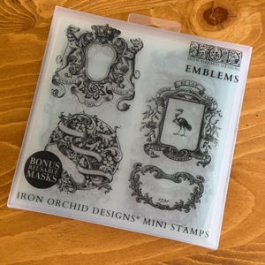May include: Clear plastic package of Iron Orchid Designs mini stamps. The stamps feature ornate emblem designs, including crests, frames, and a circular design with ribbons. The package includes the text "Emblems" and "Bonus Reusable Masks".