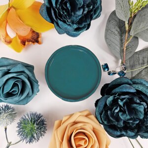 Op de afbeelding: Een tealblauwe verfstaal in een ronde container, omringd door verschillende bloemen en groen. De bloemen omvatten een gele orchidee, een tealblauwe roos en een perzikkleurige roos.