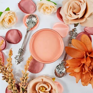 Puede incluir: Primer plano de un tarro de vela rosa con dos cucharas decorativas plateadas. El tarro está rodeado de pétalos de flores rosas y naranjas.