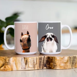 K&ouml;nnte beinhalten: Zwei wei&szlig;e Keramikbecher. Ein Becher zeigt ein Pferdgesicht mit dem Namen "Dash" dar&uuml;ber. Der andere Becher hat ein schwarz-wei&szlig;es Kaninchengesicht mit dem Namen "Oreo" dar&uuml;ber. Beide Becher haben einen wei&szlig;en Griff und stehen auf einer Holzoberfl&auml;che.