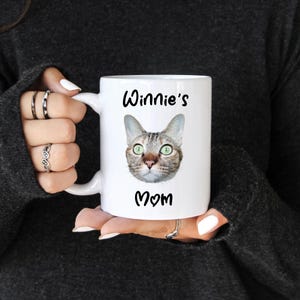 K&ouml;nnte beinhalten: Wei&szlig;e Keramik-Tasse mit einem Katzenkopf mit gr&uuml;nen Augen. Auf der Tasse steht in schwarzer Schrift der Text "Winnie's Mom". Die Tasse wird von einer Person gehalten, die einen dunkelgrauen Pullover tr&auml;gt.