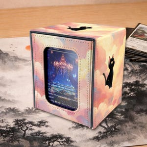 May include: A card storage box with a pastel sky design and a black cat silhouette. The box has a clear window displaying a card. Additional cards are visible on the surface.