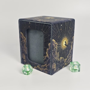 Peut inclure: Une boîte de jeu rectangulaire ornée d'un imprimé céleste bleu foncé et doré représentant des nuages, des étoiles et une main tendant vers un soleil brillant. La boîte possède une découpe centrale. Deux dés polyédriques verts sont posés devant.