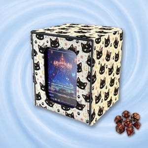 May include: A square deck box with a cream-colored exterior featuring a repeating pattern of black cat faces, small pink stars, and yellow crescent moons. A central clear window displays a game card. Seven multi-colored polyhedral dice sit nearby.