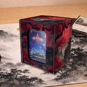 May include: A cube-shaped card storage box with a red and black floral design. The box has a window displaying a card with a fantasy landscape. The box is on a playmat with a landscape design.