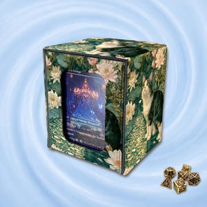 May include: A square deck box featuring a green botanical print with white water lilies and a cat, with a central window displaying a trading card. Beside the box are several small, multi-sided gold-colored dice with black numbering.