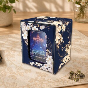 May include: A navy blue card storage box with a floral design. White flowers and gold accents adorn the box, which has a clear window displaying a card. Dice are visible in the foreground.