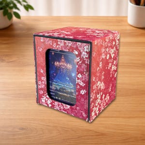 May include: A red card storage box with a floral pattern. The box has a clear window displaying a card with an image of a chandelier. The box is sitting on a wooden surface.