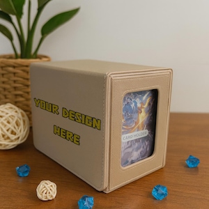 May include: A tan card holder box with the text "YOUR DESIGN HERE" in gold. The box has a window displaying a card. Decorative blue gems and a woven ball are on the wooden surface.