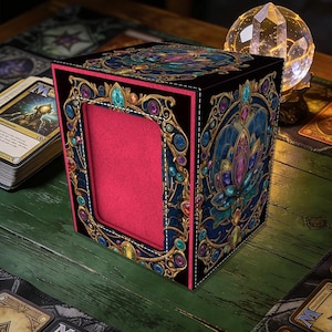Op de afbeelding: Een zwarte kaartenopbergdoos met een decoratief ontwerp met een lotusbloem en kleurrijke edelstenen. De doos heeft een rode binnenkant en gouden rand. De doos staat op een groene houten ondergrond.