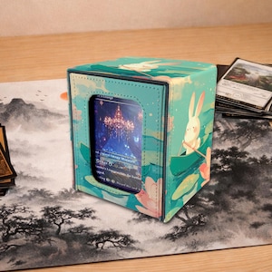 Puede incluir: Una caja de almacenamiento de tarjetas de color turquesa con un diseño caprichoso que presenta un conejo en un bote. La caja tiene una ventana transparente que muestra una tarjeta con una escena de paisaje. La caja está sobre una alfombra de juego con un diseño de paisaje.