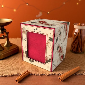 May include: A decorative box with a red interior and a window. The box is adorned with a koi fish and floral pattern. The box is on a burlap surface with cinnamon sticks.
