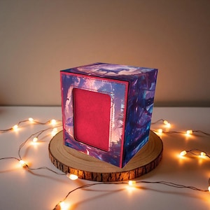 May include: A decorative cube-shaped box with a red interior and a window. The exterior features a colorful, artistic design with shades of blue, purple, and pink. The box sits on a wooden slice, with string lights around it.