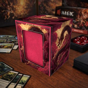 May include: A red and gold card storage box with a dragon design. The box has a rectangular opening and is decorated with a dragon and cloud motif. The box is sitting on a wooden surface with playing cards.