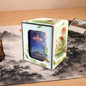 May include: A white card storage box with a window displaying a card. The box features a frog illustration with a red hat and a lotus flower design. The box is on a playmat with a landscape design, and there are other cards nearby.