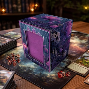 May include: A cube-shaped card storage box with a purple interior and a floral design. The exterior features a dark blue background with purple lotus flowers and skull-like imagery. White stitching outlines the edges.
