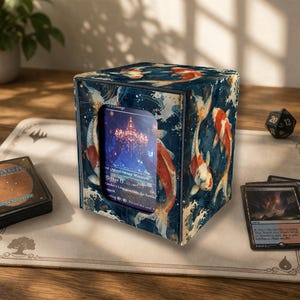 Può includere: Una scatola quadrata per riporre carte con un motivo di pesci koi blu. La scatola ha una finestra trasparente che mostra una carta con un'immagine colorata. Un mazzo di carte e un dado sono visibili.