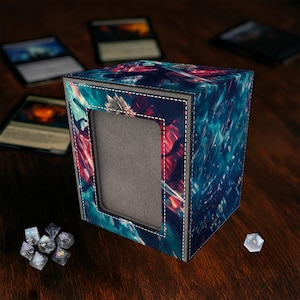 May include: A cube-shaped card storage box with a colorful abstract design in shades of blue, red, and white. The box has a rectangular opening and is surrounded by a white stitched border. Dice and cards are visible on a wooden surface.