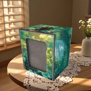 Op de afbeelding: Vierkante tissuebox met een natuurthema in groen- en geeltinten. De doos heeft een rechthoekige opening en witte stiksels. Hij staat op een ronde houten tafel.