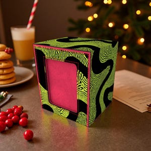 Könnte beinhalten: Eine dekorative, würfelförmige Box mit einem lebendigen Design. Die Box hat ein schwarzes und neongrünes Wirbelmuster, einen leuchtend rosa Innenraum und einen rosa Rand. Die Box steht auf einem Tisch mit einem Getränk und Keksen.
