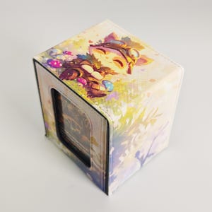 Peut inclure: Une boîte de jeu rectangulaire ornée d'une illustration colorée représentant un personnage ressemblant à un renard avec des lunettes, entouré de petits champignons. La boîte a une surface texturée et une découpe rectangulaire centrale.