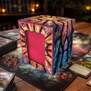 May include: A multi-colored, stained-glass-style card storage box with a red interior. The box is rectangular with a large opening and is decorated with a variety of colors. Several red dice are scattered around the box on a play mat.