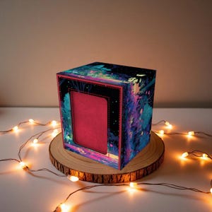 Peut inclure: Une boîte décorative ornée d'une scène de forêt vibrante dans des tons de bleu, violet et rose. L'intérieur de la boîte est rouge et elle est présentée sur une tranche de bois, avec des guirlandes lumineuses.