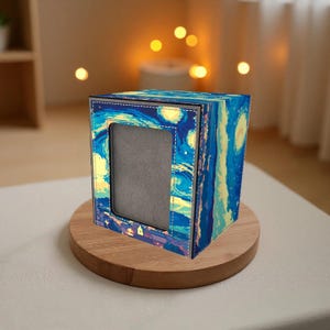 Puede incluir: Una caja cuadrada con una abertura rectangular, con una impresión de "La noche estrellada" de Van Gogh. La caja es azul, amarilla y blanca, con un borde cosido. Se encuentra sobre una base de madera redonda.
