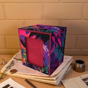 Op de afbeelding: Een vierkante doos met een levendig tropisch bladerpatroon in roze, paars, teal en zwart. De doos heeft een rechthoekige opening aan één kant, die een roze interieur onthult. Het staat op een stapel papier.