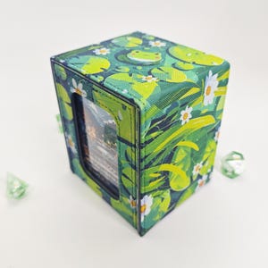 May include: A rectangular deck box featuring a vibrant green pond pattern with lily pads, white flowers, and a small frog illustration. The front panel has a clear window displaying a card, and two translucent green polyhedral dice sit nearby.