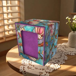 Op de afbeelding: Een decoratieve tissuebox met een glas-in-lood bloemenontwerp. De doos heeft een levendige mix van blauwe, roze en gouden kleuren, met een paars interieur zichtbaar door de opening. De doos staat op een ronde houten tafel met een kanten kleedje.