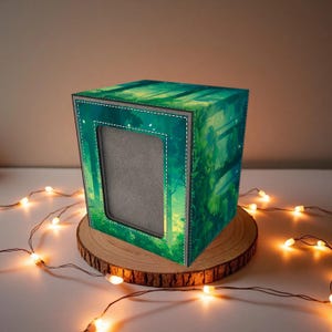 May include: A square photo frame with a forest scene design. The frame is green with a gray border and a white stitched outline. It sits on a wooden slice with string lights.