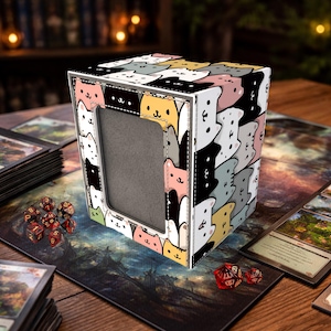 May include: A cube-shaped card storage box featuring a repeating pattern of cartoon cats in various colors. The box has a rectangular opening on one side and is surrounded by playing cards and dice on a wooden table.