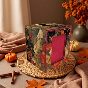 May include: A floral-patterned tissue box with a dark background and red, orange, and cream-colored flowers. The box has a rectangular opening and a red interior. It sits on a woven mat with pumpkins and fall leaves.