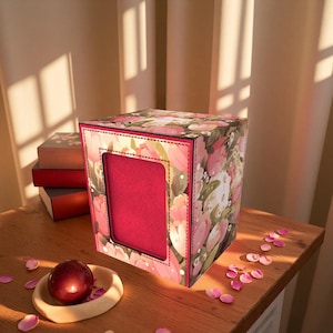 May include: A floral-patterned box with a rectangular opening, featuring pink and white flowers on a dark background. The box sits on a wooden surface with a red sphere in a wooden bowl and scattered pink petals. Books are visible in the background.
