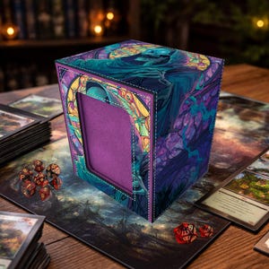 May include: A cube-shaped card storage box with a stained-glass design featuring a hooded figure in shades of blue, purple, and teal. The box has a purple interior and is surrounded by playing cards and dice. The box is approximately 4 inches tall.